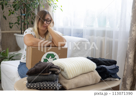 A thoughtful woman sits near a box with neatly folded clothes. She sorts her clothes for recycling or donation, emphasizing the importance of clothing recycling. Recycling, environmental awareness. A thoughtful woman sits near a box with neatly folded clothes. She sorts her clothes for recycling or donation, emphasizing the importance of clothing recycling. Recycling, environmental awareness. 113071390