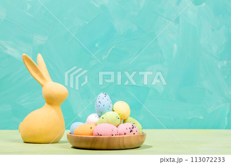 Easter card. Yellow bunny and decorative colorful eggs in wooden plate on turquoise Copy space. 113072233
