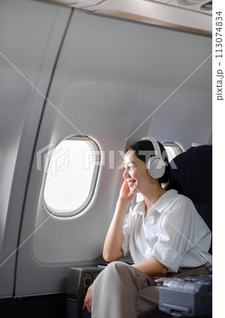 Asian woman sitting and using a smartphone in an airplane Listening to music next to the airplane seat window travel and technology concept 113074834