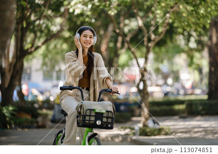 Young asian woman using headphones while riding bicycle in city park Young asian woman using headphones while riding bicycle in city park 113074851