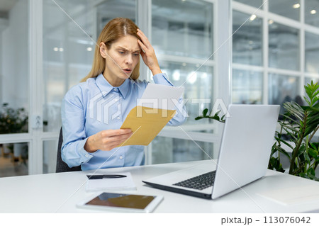 A businesswoman in a blue shirt appears perplexed analyzing documents beside her laptop in a bright office setting. 113076042
