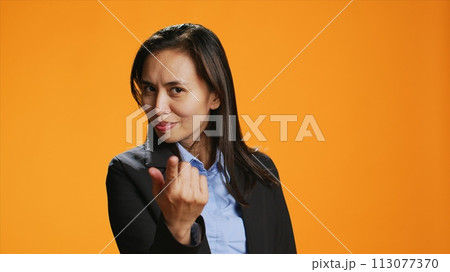 Female model calling for someone on camera, doing hailing gesture against orange background. Asian woman with office clothing inviting person to come closer, friendly adult. 113077370