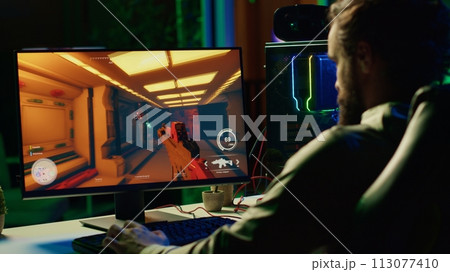 Man in dark living room playing engaging video games on gaming PC at computer desk, chilling after work. Gamer contending against foes in online multiplayer shooter, shooting them with lasers 113077410