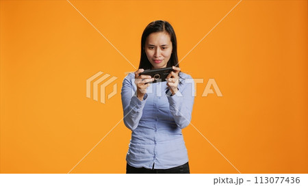 Joyful adult playing mobile videogames on camera, having fun with modern shooting competition online on smartphone network. Asian woman using phone for entertainment and leisure. Joyful adult playing mobile videogames on camera, having fun with modern shooting competition online on smartphone network. Asian woman using phone for entertainment and leisure. 113077436