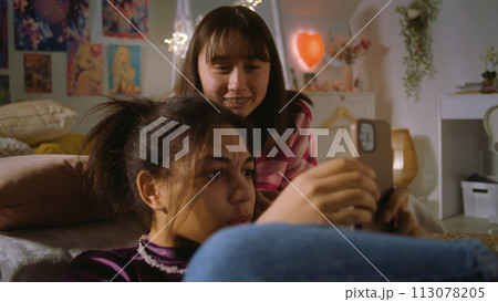 Asian teen lies on bed and watches content with friend 113078205