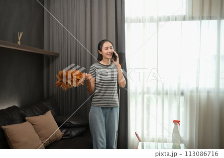Pretty young housewife with dust brush talking on mobile phone in living room 113086716