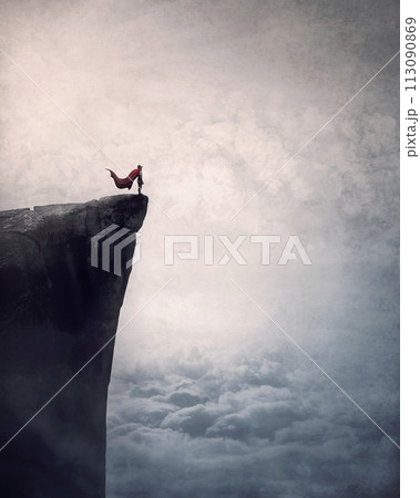 Superhero with cape stands brave on edge of a cliff above the clouds searching determined at horizon with hand up to forehead. Powerful hero conceptual scene 113090869