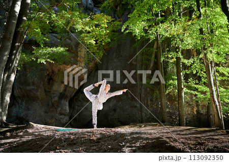 Woman practicing yoga outdoors in forest. Barefoot female on yoga mat surrounded by trees and large rocks, which suggests peaceful, natural environment ideal for meditation or yoga practice. Woman practicing yoga outdoors in forest. Barefoot female on yoga mat surrounded by trees and large rocks, which suggests peaceful, natural environment ideal for meditation or yoga practice. 113092350