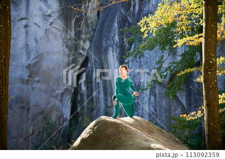Woman practicing yoga outdoors in forest. Barefoot female on yoga mat surrounded by trees and large rocks, which suggests peaceful, natural environment ideal for meditation or yoga practice. 113092359