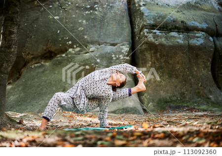 Woman practicing yoga outdoors in forest. Barefoot female on yoga mat surrounded by trees and large rocks, which suggests peaceful, natural environment ideal for meditation or yoga practice. 113092360