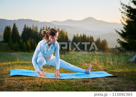 Woman practicing yoga outdoors in the mountains in a serene, natural setting. Female performing yoga pose on blue mat, with backdrop of beautiful mountain landscape at sunrise or sunset. 113092365