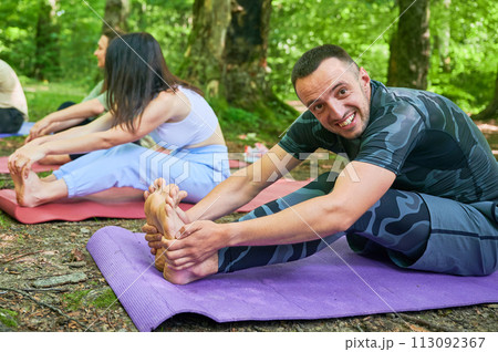 People engaged in stretching exercise outdoors, part of yoga practice. Portrait of happy man stretching forward to reach his toes, sitting on purple yoga mat. 113092367