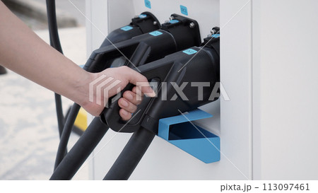 Hand holding EV charging plug in front of camera with blurred background of outdoor. Driver of electric car hold batteries charger plug. Sustainable energy powered electric charging station background 113097461