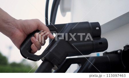 Hand holding EV charging plug in front of camera with blurred background of outdoor. Driver of electric car hold batteries charger plug. Sustainable energy powered electric charging station background Hand holding EV charging plug in front of camera with blurred background of outdoor. Driver of electric car hold batteries charger plug. Sustainable energy powered electric charging station background 113097463