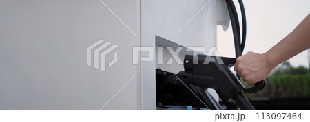 Hand holding EV charging plug in front of camera with blurred background of outdoor. Driver of electric car hold batteries charger plug. Sustainable energy powered electric charging station background 113097464