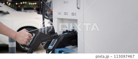 Hand holding EV charging plug in front of camera with blurred background of outdoor. Driver of electric car hold batteries charger plug. Sustainable energy powered electric charging station background 113097467