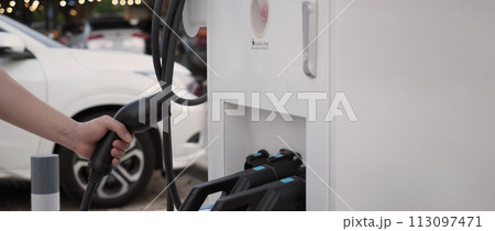 Hand holding EV charging plug in front of camera with blurred background of outdoor. Driver of electric car hold batteries charger plug. Sustainable energy powered electric charging station background 113097471