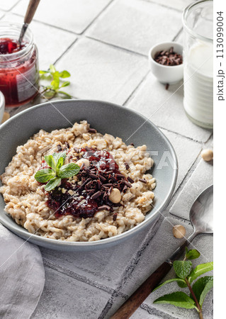 Oatmeal porridge with raspberry jam and nuts in gray bowl close up on gray tile background served with spoon. Healthy diet breakfast 113099004