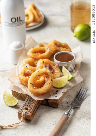 Crisp fried golden squid rings with lemon and sauce on wooden board, light background Crisp fried golden squid rings with lemon and sauce on wooden board, light background 113099018