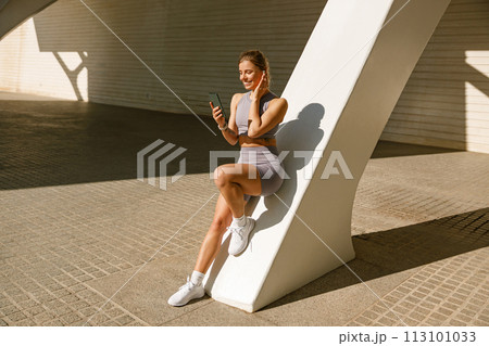 A young woman in sportswear is resting after a workout outside while standing with smartphone 113101033