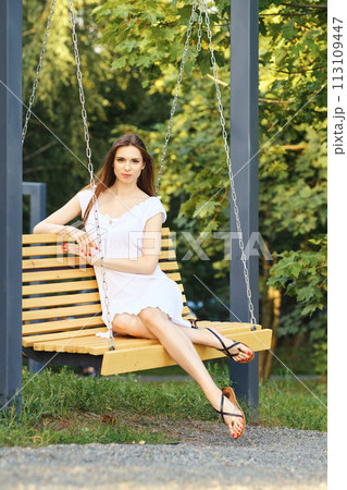 Cheerful woman in white dress enjoys swinging on a swing in city park 113109447