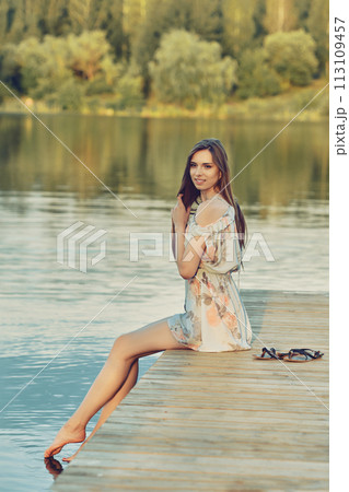 Woman sitting on the edge of dock in natural park during summer looking meditative at the water 113109457