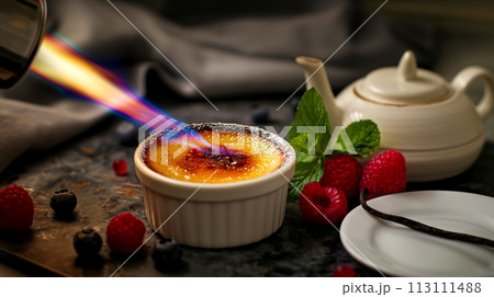 The chef burns sugar on a portion of creme brulee, the dessert is served with fresh berries and mint leaves, a popular dessert made from cream 113111488