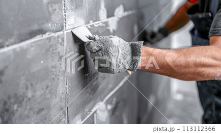 A builder specialist tiler applies a mortar with a spatula to seal the fugue on the tiles, a concept for the renovation of apartments and offices A builder specialist tiler applies a mortar with a spatula to seal the fugue on the tiles, a concept for the renovation of apartments and offices 113112366