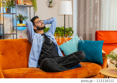 Happy relaxed Hispanic man resting on home couch with wireless earphones listening ambient music 113116112