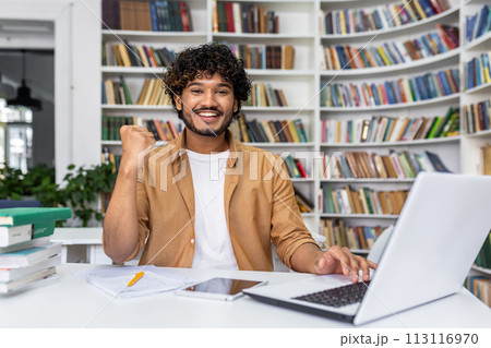 Joyful young man triumphantly clenches fist looking at laptop in library, expressing excitement for his achievement or win. 113116970
