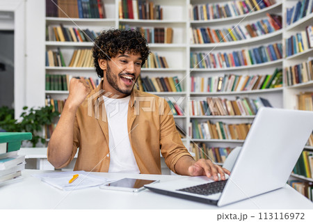 A young professional man in a casual outfit working on his laptop in a modern office environment, surrounded by books. A young professional man in a casual outfit working on his laptop in a modern office environment, surrounded by books. 113116972