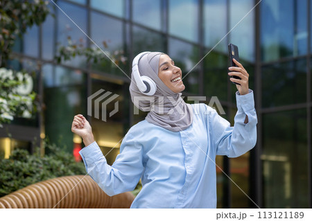 A modern Muslim woman in a hijab is capturing a selfie with her smartphone outside a reflective glass office building. 113121189