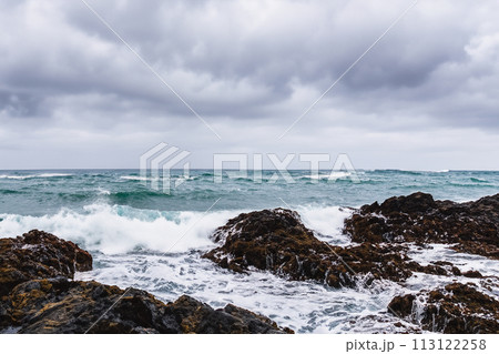 Surf on a rocky coast in front of the horizon under a cloudy sky. Tropical stormy ocean with waves crashing on the rocks. Stormy sea with stormy clouds and black lava rocks on the shore. Splashing 113122258