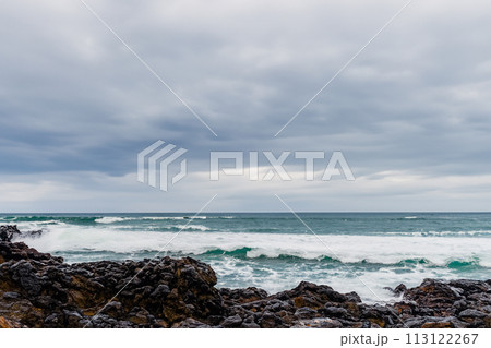 Surf on a rocky coast in front of the horizon under a cloudy sky. Tropical stormy ocean with waves crashing on the rocks. Stormy sea with stormy clouds and black lava rocks on the shore. Splashing Surf on a rocky coast in front of the horizon under a cloudy sky. Tropical stormy ocean with waves crashing on the rocks. Stormy sea with stormy clouds and black lava rocks on the shore. Splashing 113122267