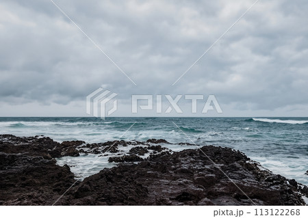 Surf on a rocky coast in front of the horizon under a cloudy sky. Tropical stormy ocean with waves crashing on the rocks. Stormy sea with stormy clouds and black lava rocks on the shore. Splashing Surf on a rocky coast in front of the horizon under a cloudy sky. Tropical stormy ocean with waves crashing on the rocks. Stormy sea with stormy clouds and black lava rocks on the shore. Splashing 113122268