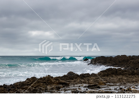 Surf on a rocky coast in front of the horizon under a cloudy sky. Tropical stormy ocean with waves crashing on the rocks. Stormy sea with stormy clouds and black lava rocks on the shore. Splashing Surf on a rocky coast in front of the horizon under a cloudy sky. Tropical stormy ocean with waves crashing on the rocks. Stormy sea with stormy clouds and black lava rocks on the shore. Splashing 113122275