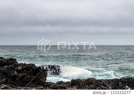 Surf on a rocky coast in front of the horizon under a cloudy sky. Tropical stormy ocean with waves crashing on the rocks. Stormy sea with stormy clouds and black lava rocks on the shore. Splashing Surf on a rocky coast in front of the horizon under a cloudy sky. Tropical stormy ocean with waves crashing on the rocks. Stormy sea with stormy clouds and black lava rocks on the shore. Splashing 113122277