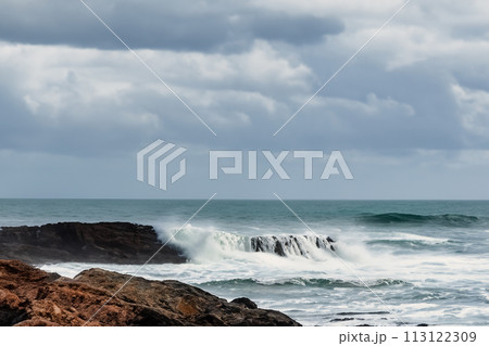 Surf on a rocky coast in front of the horizon under a cloudy sky. Tropical stormy ocean with waves crashing on the rocks. Stormy sea with stormy clouds and black lava rocks on the shore. Splashing Surf on a rocky coast in front of the horizon under a cloudy sky. Tropical stormy ocean with waves crashing on the rocks. Stormy sea with stormy clouds and black lava rocks on the shore. Splashing 113122309