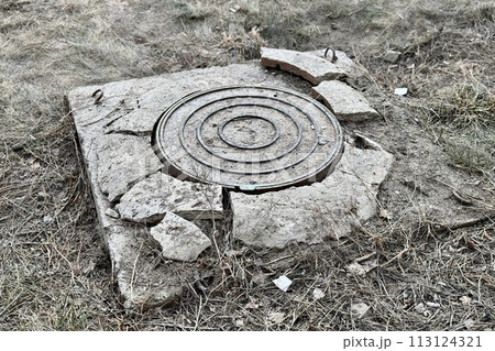 A rusty iron hatch with a pattern and a large gap at the top is mounted in a broken cracked asphalt structure well next to the road. Close-up 113124321