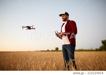 Farmer holds remote controller with his hands while quadcopter is flying on background. Drone hovers behind the agronomist in wheat field. Agricultural new technologies and innovations. Back view Farmer holds remote controller with his hands while quadcopter is flying on background. Drone hovers behind the agronomist in wheat field. Agricultural new technologies and innovations. Back view 113125814
