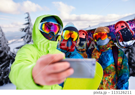 Three snowboarders taking selfie with smartphone camera at ski resort. Friends photographing for social network sharing with snowboards near forest wearing reflective goggles, colorful fashion clothes 113126211