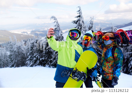 Three snowboarders taking selfie with smartphone camera at ski resort. Friends photographing for social network sharing with snowboards near forest wearing reflective goggles, colorful fashion clothes Three snowboarders taking selfie with smartphone camera at ski resort. Friends photographing for social network sharing with snowboards near forest wearing reflective goggles, colorful fashion clothes 113126212