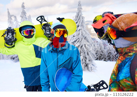 Three snowboarders walking at ski resort. Friends climbing to mountain top carrying their snowboards through forest for backcountry freeride and wearing reflective goggles, colorful fashion clothes. 113126222