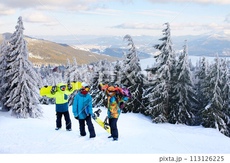Three snowboarders walking at ski resort. Friends climbing to mountain top carrying their snowboards through forest for backcountry freeride and wearing reflective goggles, colorful fashion clothes. Three snowboarders walking at ski resort. Friends climbing to mountain top carrying their snowboards through forest for backcountry freeride and wearing reflective goggles, colorful fashion clothes. 113126225