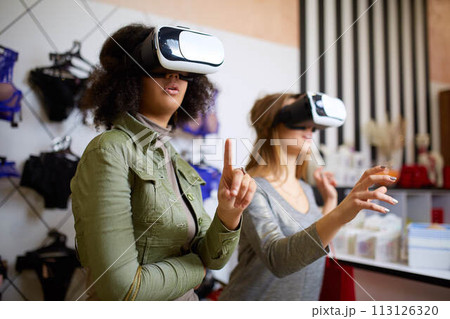 Two women in modern virtual reality headsets having expirience in shopping at lingerie store. Multiracial girls in vr glasses with bags touching and pointing interface elements in underwear shop. Two women in modern virtual reality headsets having expirience in shopping at lingerie store. Multiracial girls in vr glasses with bags touching and pointing interface elements in underwear shop. 113126320
