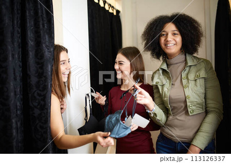 Beautiful young woman asking for a friends opinion in the fitting room of lingerie store. Female tries on bra in underwear shop. Shop assistant helps girl to choose right size in changing room. Beautiful young woman asking for a friends opinion in the fitting room of lingerie store. Female tries on bra in underwear shop. Shop assistant helps girl to choose right size in changing room. 113126337