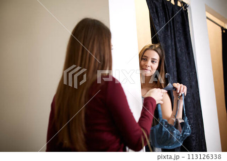 Beautiful young woman asking for a friends opinion in the fitting room of lingerie store. Female tries on bra in underwear shop. Shop assistant helps girl to choose right size in changing room. 113126338