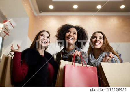 Women of diverse ethnicity with shopping bags posing in mall on sale. Portrait of three smiling multiracial girls look up at the copyspace area above their heads for designer. 113126421