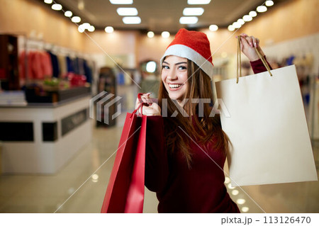 Happy girl shopping gifts in mall on christmas sale. New year holidays shopping idea concept. Smiling pretty caucasian woman with colorful paper presents bags wearing christmas hat in store or shop. 113126470