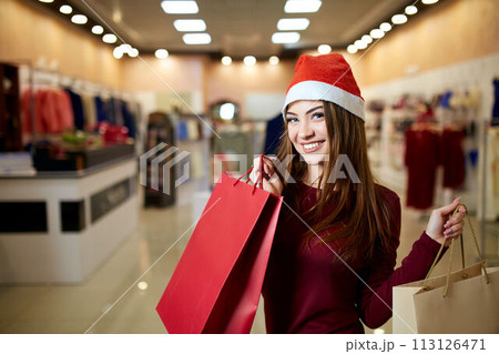 Happy girl shopping gifts in mall on christmas sale. New year holidays shopping idea concept. Smiling pretty caucasian woman with colorful paper presents bags wearing christmas hat in store or shop. Happy girl shopping gifts in mall on christmas sale. New year holidays shopping idea concept. Smiling pretty caucasian woman with colorful paper presents bags wearing christmas hat in store or shop. 113126471